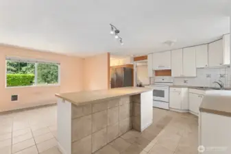 This view shows how the kitchen stays bright and airy, with light bouncing off white finishes and oversized windows. The bar counter is finished with protective tile underneath, so stool scuffs never stand a chance. Across from the island, another cadet wall heater lets you dial in the temperature room by room, and blinds are already in place when you want to control the light.