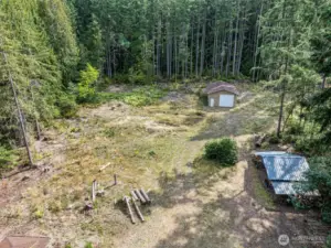 Aerial of partially cleared property, nearly level and lined with trees for privacy. Oversized shop/garages towards the back treeline. Carport on the right and outbuilding with additional storage and well pressure tank on the lower left.
