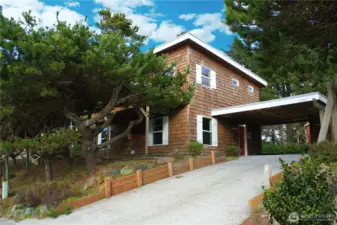 A cedar-shingled coastal home with a covered carport and a tree-lined concert driveway.