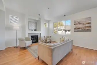 Bright and inviting living room features a cozy fireplace framed by decorative detailing and flanked by charming accent windows that bring in soft natural light. A large picture window enhances the airy feel while offering neighborhood views. Thoughtful furnishings, warm wood flooring, and a clean, contemporary palette create a comfortable and elegant space ideal for everyday living and relaxed entertaining.