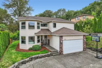 This two-story home features a modern exterior with clean lines and a welcoming front entrance. Large, bay-style windows on the front add a sophisticated touch, allowing for ample natural light inside while enhancing the home's curb appeal. The exterior is a blend of light siding and brick accents, adding a charming contrast and classic style. A spacious driveway leads to a two-car garage, perfect for convenience and storage. The landscaped front yard is neatly designed with shrubs and a stone-lined garden bed. A basketball hoop in the driveway adds a playful, family-friendly element. The house is nestled in a tranquil, tree-lined neighborhood, offering privacy and scenic surroundings.