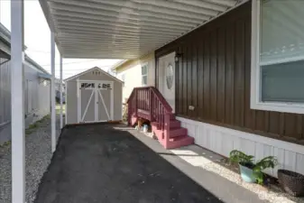 This home features a practical and covered outdoor area, offering protection from the elements year-round. The space provides easy access to a dedicated storage shed, perfect for keeping tools, bikes, and other outdoor gear organized and out of sight. A set of steps with a handrail leads to the home's entrance, providing both accessibility and a welcoming feel. This functional design makes outdoor living simple and tidy.