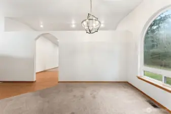 The formal dining room keeps its own identity while still feeling connected to the rest of the house. A large arched window brings in soft natural light, with operable lower panes for fresh air, and the contemporary chandelier centers the space nicely. The partial height wall and arched opening into the kitchen create separation for meals and conversations while still allowing light and airflow to pass through. From here you can see the hardwood flooring in the hall leading toward the kitchen, tying the rooms together visually even as the dining area holds its quieter feel.