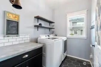 Unique laundry room, dark grey ceramic ties, cement counter top, subway backsplash, heavy wood and iron shelving, vented window, crown molding and dark wood tone cabinet.  The entry door to this spacious room is an antique white washed wood & glass door with window pane cut outs.  Washer & dryer stay with the home. Wall lighting is for decor purposes only, non functional.