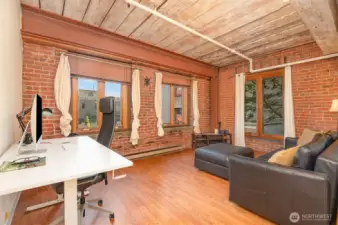 Experience versatile living in this spacious 815 sq ft condo, featuring a stylish living area with warm hardwood floors and exposed brick walls. The photo highlights a modern workspace  complemented by large windows with natural light. Exposed wooden beams and steel accents add historic charm, making this Seattle waterfront retreat perfect for work and relaxation!