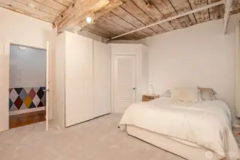 Unwind in this serene 1-bedroom retreat, featuring a plush carpeted floor and a cozy bed with soft linens. The photo highlights exposed brick walls and wooden beams, adding historic charm, while large windows with A/C offer natural light and comfort.