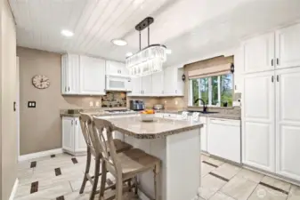 The revamped kitchen includes an island with seating, ceramic tile flooring, white cabinetry with tons of storage, granite countertops, and a gas cooktop.