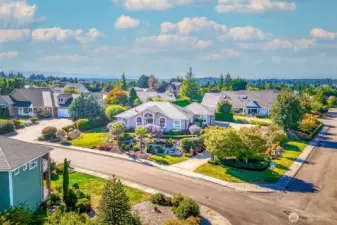 Striking aerial view showcases this elegant single-level home on a beautifully landscaped corner lot. Lush gardens, palms, and manicured beds frame the residence, creating exceptional curb appeal in a desirable neighborhood.