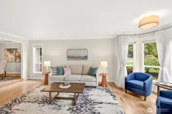 The formal living room is filled with natural light from the bay window, complemented by custom blinds and a clean, modern overhead fixture, all anchored by rich hardwood flooring.