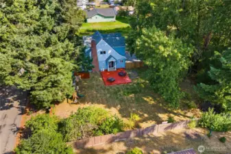 Angled aerial view showing the full backyard, red deck, and fenced perimeter, framed by tall evergreens. A great vantage point for the size of the lot and outdoor living space.
