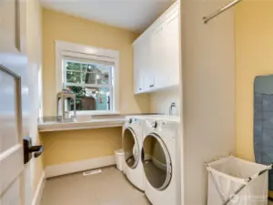Everyday tasks feel easier in this main-floor laundry, complete with a folding counter, drying bar, generous cabinetry, and tile floors. A sunny window brings in natural light for a bright, efficient workspace.