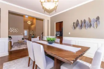 Elegant dining room with wainscoting and crown molding, open to the foyer and kitchen for effortless entertaining. A neutral palette and natural light highlight the rich wood floors, while metal leaf wall art adds a modern touch.