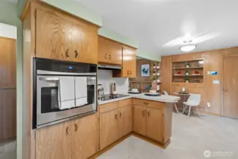 Built-in oven and cooktop are framed by classic cabinetry, creating a dedicated cooking zone. The open pass-through connects the kitchen to the adjacent dining nook with built-in shelving nearby.