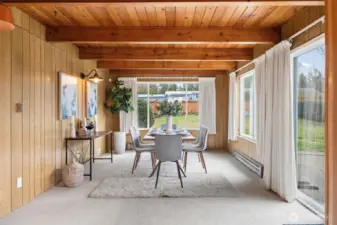 Dining area features warm wood paneling and exposed ceiling beams that add architectural character to the space. Large windows and a sliding glass door frame backyard views while bringing in abundant natural light for everyday gatherings.
