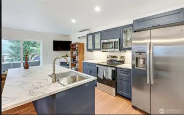 Kitchen of DADU featuring an island with overhang slab offering extra counter space and bar seating area, large stainless side-by-side fridge, and matching stainless appliances including a glass cook top range/oven and above-mount microwave. Upper display cabinets to show off your dishes or staple pieces. Mini-split located right of kitchen in the living room area near the electric fireplace.