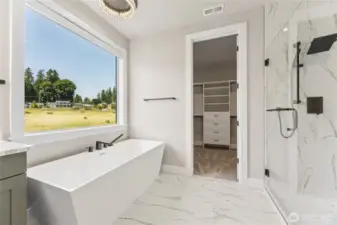 This stunning primary bath features a spa-inspired soaking tub with peaceful views, marble-style tile flooring, a glass-enclosed shower with matte black fixtures, and direct access to a spacious walk-in closet.