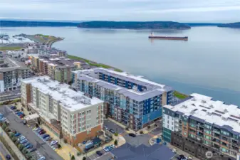 A sweeping aerial view showcases the Baker Building along the shores of Commencement Bay, highlighting its exceptional location within the vibrant Point Ruston waterfront community. Enjoy immediate access to shoreline trails, seaside dining, and year-round outdoor recreation.