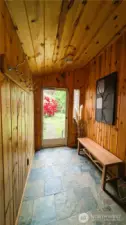 This charming wood-paneled foyer blends rustic textures with practical design, featuring stone tile flooring, vintage hardware, and warm natural light that flows in through the glass door beyond.