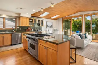A beautifully appointed kitchen where rich wood cabinetry and warm hardwood floors surround a professional-grade Viking gas 6-burner range, creating a chef's space that flows effortlessly into the family room and out toward the sun-drenched deck beyond.