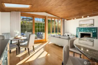 A seamless indoor-outdoor living space where the striking slatted wood ceiling, gleaming hardwood floors, and a cozy fireplace are bathed in natural light pouring through expansive glass doors — opening directly onto a lush deck and inviting the outdoors in at every turn.
