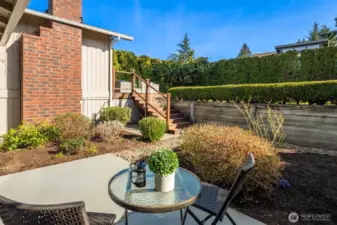 A private east-facing patio retreat just off the primary bedroom, bathed in glorious morning sun, where a charming bistro seating area nestles against classic brick and mature landscaping — a peaceful outdoor escape perfect for morning coffee or quiet evening unwinding.