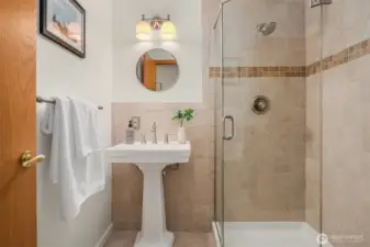 A clean and classic lower level ¾ bathroom featuring a elegant pedestal sink, glass-enclosed shower with warm tile surround, and timeless finishes — a well-appointed space that completes the lower level bedroom suite and enhances the home's mother-in-law conversion potential.