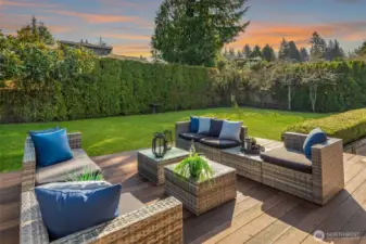 A beautifully manicured backyard oasis featuring a generous deck with stylish wicker lounge seating, opening onto a pristine, flat lawn framed by mature trees and lush landscaping — a private, sun-drenched retreat perfectly designed for entertaining and outdoor living at its finest.