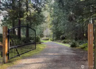 A gravel driveway stretches past the gate, winding gently through peaceful acreage beyond. The land feels expansive and secluded, offering privacy and tranquility.
