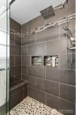 Luxury walk-in shower at 4612 N 21st St, Tacoma, WA — featuring large-format modern tile, built-in shelving niches, mosaic tile flooring, a rainfall showerhead, handheld sprayer, and a built-in bench for comfort. This North Tacoma home offers a beautifully upgraded bathroom with spa-like finishes and contemporary design.