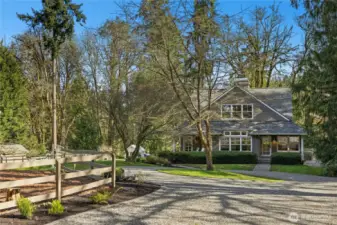 A sweeping circular drive curves to the Craftsman home, framed by mature evergreens and a peaceful, park-like setting. To the left, fenced paddock supports an equestrian lifestyle, with the greenhouse tucked in the distance for year-round growing and homestead charm.