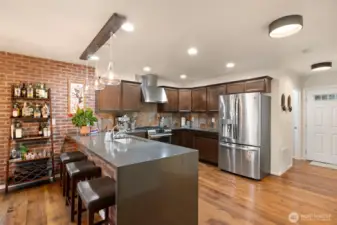 This second view of the kitchen captures the home’s welcoming flow back toward the entry. Sleek stainless-steel appliances, rich cabinetry, and quartz-style solid-surface counters set a refined tone, while custom brick veneer detailing adds warmth and character. Conveniently located just off the entry, the laundry room offers added functionality without sacrificing style — a thoughtful touch for busy everyday living