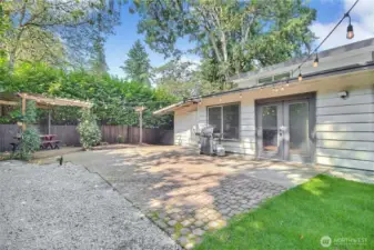 This inviting backyard features a spacious patio with string lighting, perfect for outdoor dining and entertaining. A pergola-covered seating area adds charm and shade, while the open paver space is ideal for barbecues or gatherings. Surrounded by lush greenery and a private fence, it offers both relaxation and privacy in a beautiful outdoor setting.