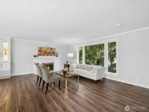Looking from foyer back through living room at main floor fireplace & large window looking out onto manicured garden beds and mature trees