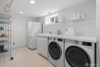 Bright and spacious laundry room with side-by-side washer/dryer, utility sink, folding counter, shelving, and bonus refrigerator—designed for efficiency and extra storage.