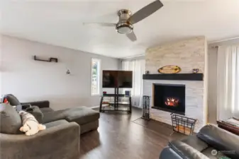 The great room features a fireplace with white brick surround, laminate wood flooring and ceiling fan.