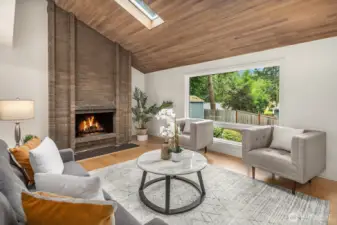 The living room's soaring vaulted ceiling, wood-planked skylight, and striking floor-to-ceiling fireplace combine to create a warm and architectural centerpiece that is truly one of a kind. Large windows frame the lush greenery outside, flooding this beautifully updated space with natural light and a seamless connection to the outdoors.