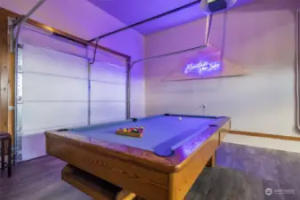 Garage game room with custom neon sign, professional grade pool table, ping pong, foosball, LVP flooring throughout, vehicle access at front and back of the garage.