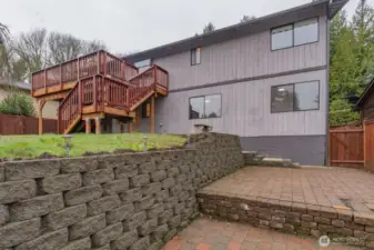 The deck provides easy access to the terraced backyard featuring a professionally installed interlocking concrete block retaining wall, creating a level patio and usable outdoor living space ideal for entertaining, gardening, or relaxing outdoors.
