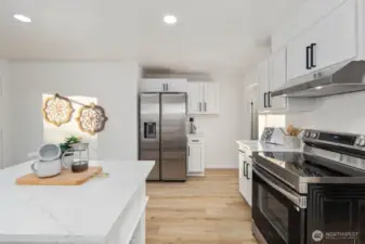 Alternate angle highlighting stainless steel refrigerator, smooth-top range with vent hood, and light wood flooring throughout.
