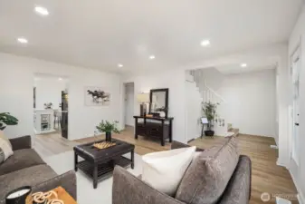 View of the living area connecting to the kitchen and staircase, featuring recessed lighting, light wood floors, and an open, inviting flow.