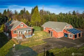 An inviting view of the main home paired with a versatile detached garage that truly adds to the lifestyle here. The garage is well set up with updated electrical, its own water heater, hot and cold water, bright LED lighting, and a spacious loft above for storage or projects. An attached greenhouse and generous paved area complete the scene, offering space to tinker, grow, and spread out while enjoying the privacy of the surrounding grounds.