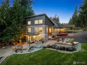 This closer aerial view gives you the full tour without ever stepping out of the drone. The wide asphalt drive leads right to the spacious covered entry, flanked by clean, low-maintenance landscaping and two separate lawn areas, and yes, there’s room for a trampoline and a cornhole set. The home’s contemporary exterior brings the curb appeal, while the elevated deck off the main floor offers front-row lake views. Below, the patio outside the Airbnb unit is perfect for guests or gatherings, and the built-in fire pit is ready to host your next marshmallow moment with the whole crew.