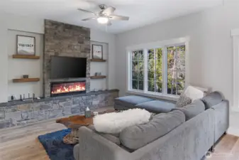 Comfortable living room featuring a beautiful stone fireplace, modern floating shelves, and expansive windows overlooking the wooded UW Preserve—perfect for cozy evenings on San Juan Island.