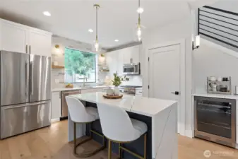 This kitchen blends elegance and efficiency with a marble-topped island, luxe gold-accented seating, and a black-railed staircase that frames the space—while the wine cooler, coffee station, and natural light from the window add everyday indulgence to its polished functionality.
