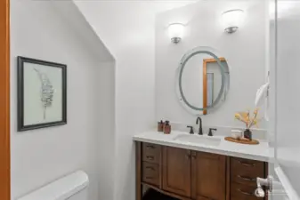 Powder room with single-sink vanity, wood cabinetry, and solid surface countertop. Round mirror and wall-mounted lighting provide a clean, functional space.