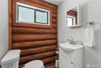Rustic-Modern Half Bath with Log Wall Accent  This charming half bath blends modern touches with rustic appeal, featuring a beautiful log wall accent that brings warm cabin character to the space. A sleek white vanity, updated fixtures & mosaic backsplash add a clean, fresh feel, while the frosted window offers natural light with privacy. Perfectly placed for guests or daily convenience, this bathroom adds style & function to your Pacific Northwest retreat.