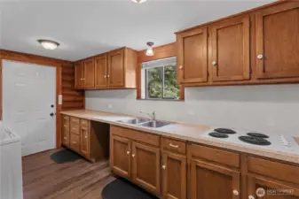 Spacious Utility Kitchen with Wood Cabinets & Exterior Access  This versatile second kitchen or utility space features abundant wood cabinetry, a dual basin sink, and built-in cooktop—perfect for canning, entertaining prep, or multi-generational living. A window above the sink offers views of the wooded surroundings, while direct access to the exterior makes this space ideal for backyard BBQ prep or garden-to-table convenience. With durable finishes & rustic charm, it adds both function & flexibility to this country property.