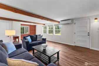 Bright Living Room with Log Cabin Accent & Beam Ceilings  This cozy living room is filled with natural light from three large windows that offer tranquil views of the surrounding acreage. Exposed wood beams and log-style accent walls add character and a warm cabin feel, while the updated flooring and mini-split AC ensure year-round comfort. With room to gather, relax, and unwind, this space blends rustic charm with modern convenience—ideal for embracing the peaceful Pacific Northwest lifestyle.