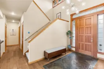 Right when you walk in, you can tell this home has presence. The eight panel solid wood door is framed with wood wrapped trim and flanked by tall privacy windows, plus that long accent window above that brings in light without putting your life on display. The slate detail at the entry handles wet shoes like a pro, then transitions into those rich maple floors that run through the main level. The space opens up to a full two story foyer with a clean, modern chandelier that keeps it feeling current. There’s room for a bench setup, which just makes everyday comings and goings easier. You’ve got quick access to the powder room, a hallway leading to the main floor suite, and a large under stair storage area that’s way more useful than expected.