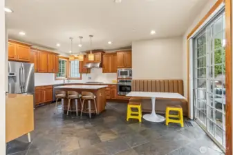 From here, the kitchen shows off its personality. The island pulls people in with casual seating, the slate floors add texture and durability, and the stainless appliances keep things current. The booth nook by the sliding door feels like a built in favorite spot, tucked right where the light comes in and close to the backyard for easy in and out. The corner sink with those two windows is one of those features you don’t realize you love until you have it, bright, open, and a little more interesting than the usual setup. Solid Cherrywood cabinets warm up the space, and everything flows naturally back into the living area.